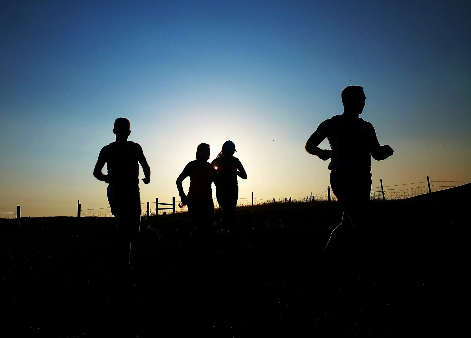 The image shows four people running outdoors. It's a silhouette, captured against a bright sky background, probably during sunrise or sunset, giving a dramatic effect to the scene.