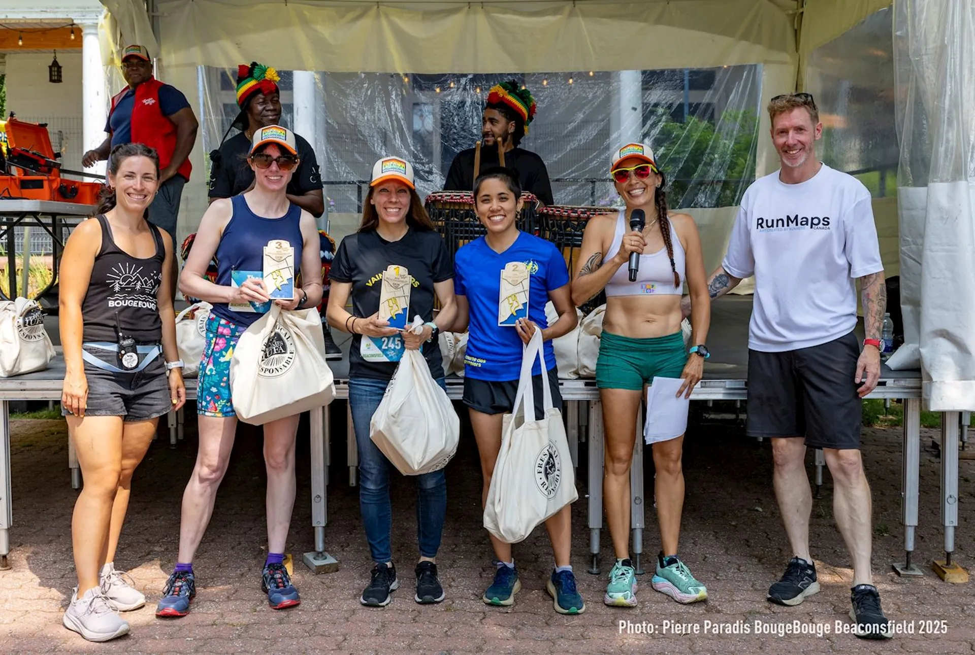 The image shows a group of people standing together, likely at an outdoor event or race. Three women in the center are holding plaques and gift bags, possibly indicating they are winners or participants in the event. There are musicians playing instruments in the background under a tent, and other people are standing nearby. The atmosphere looks celebratory.