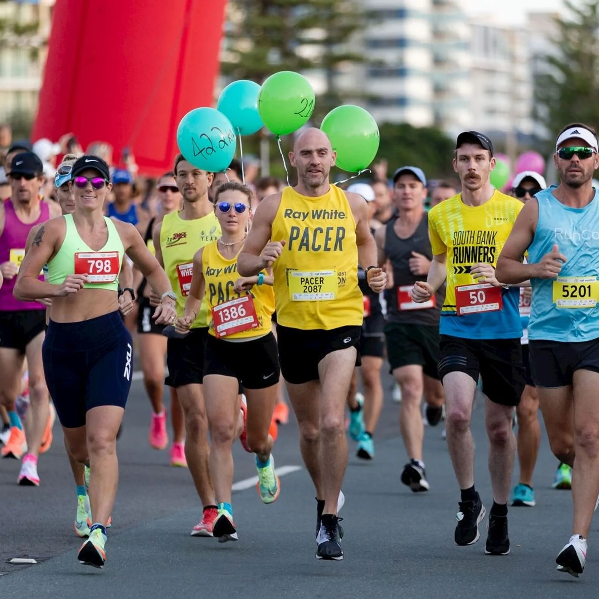 The image shows a group of runners participating in a race. One runner in the center is wearing a "Pacer" shirt and holding green balloons, which typically indicates they are setting a specific pace for other runners. There are race numbers visible on the participants' outfits, and several runners are wearing sunglasses and athletic gear. The setting appears to be an organized marathon or running event.