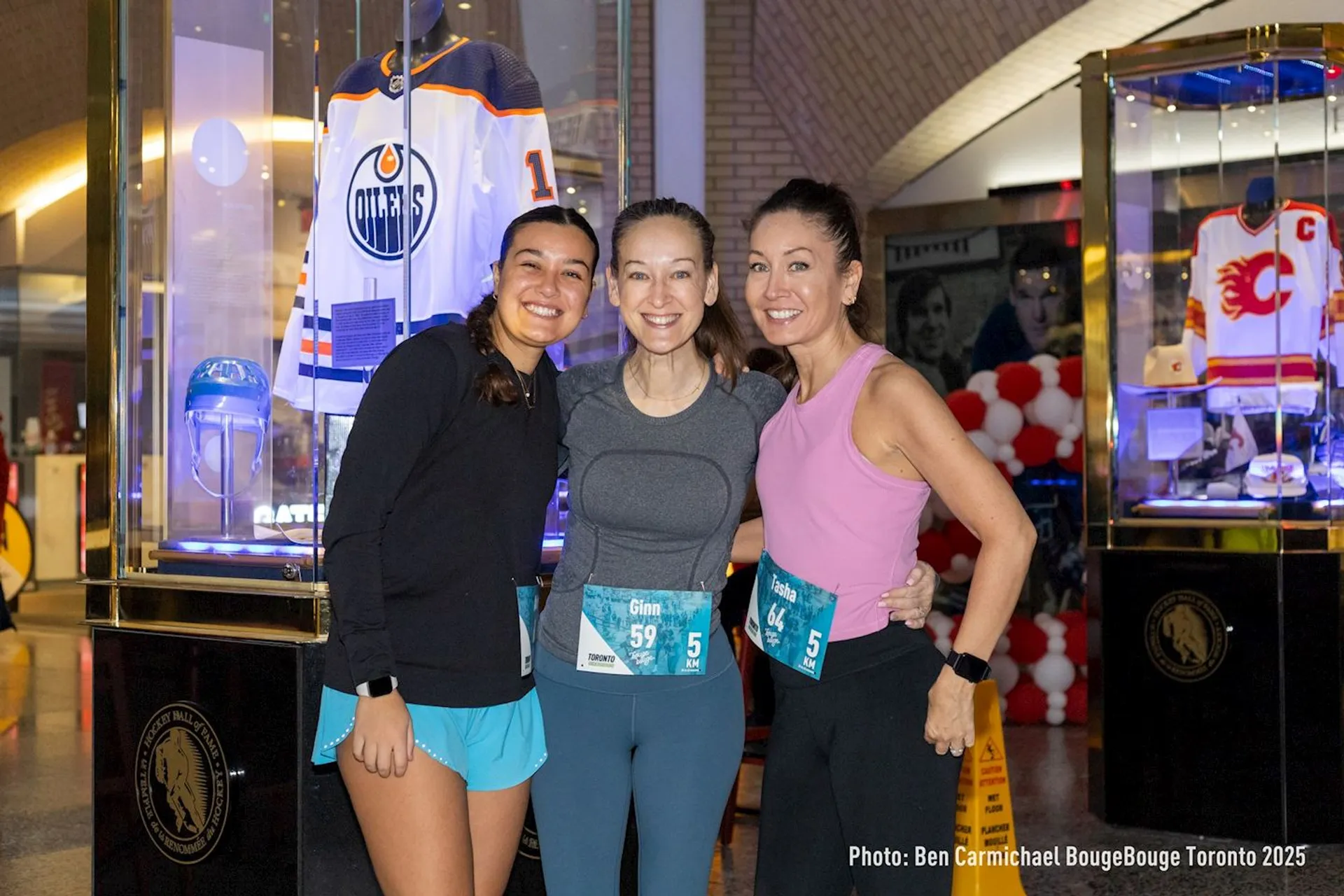 The image shows three people posing and smiling for a photo in front of display cases containing sports jerseys. The case on the left has an Edmonton Oilers jersey, and the case on the right has a Calgary Flames jersey. There are event race numbers on the people, suggesting they might be participating in a running or sporting event.