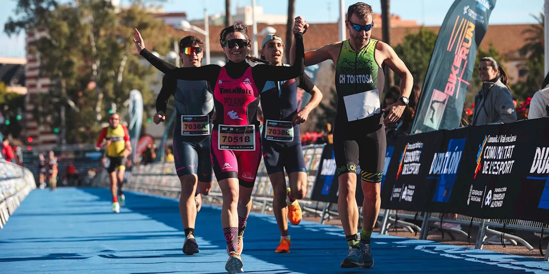 This image shows three athletes crossing the finish line at a triathlon event. They appear to be celebrating their achievement, with one of them raising their arms in the air triumphantly. They are wearing typical triathlon attire, which includes fitted tops and shorts suitable for swimming, biking, and running. Their race numbers are visible, and one can see timing chips strapped to their ankles, which are used to record their race times. There are event barriers on either side of the runners that are branded with the event's logos, and spectators can be seen in the background cheering the participants. The setting appears to be in a well-lit outdoor area with blue skies, and the overall atmosphere seems to be one of accomplishment and festivity.