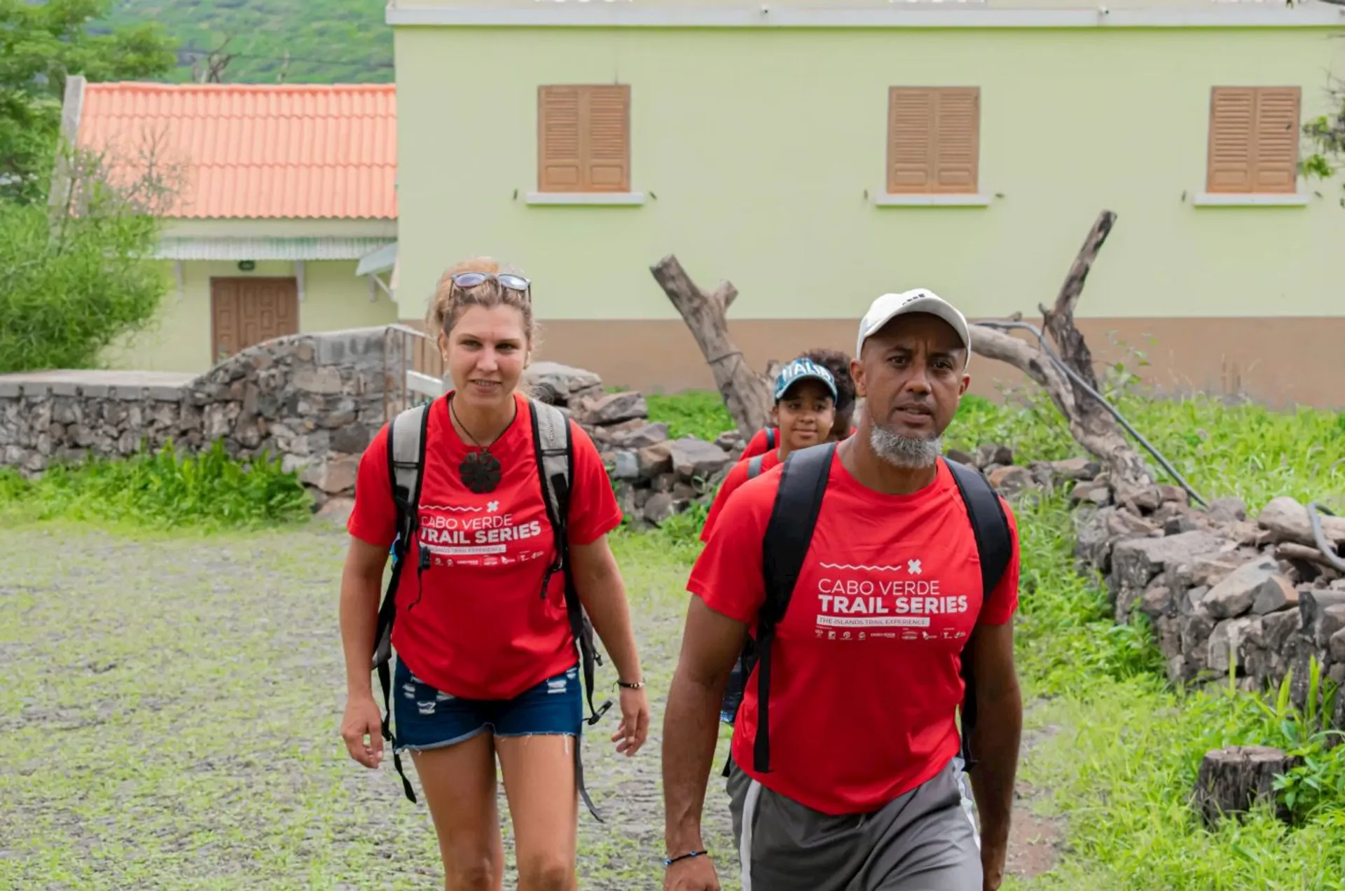 The image shows a group of people walking outdoors. They are wearing red shirts with the text "Cabo Verde Trail Series." The background includes a building, some trees, and a stone wall.