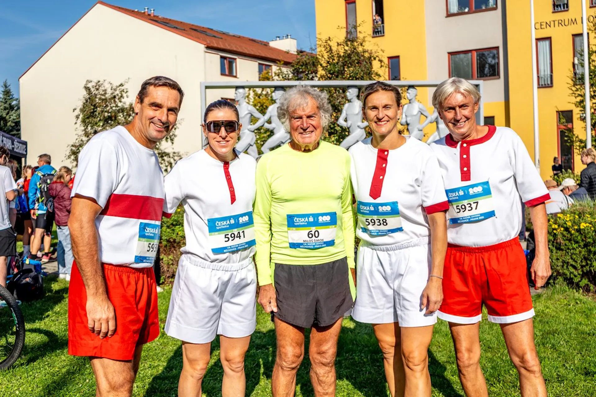 The image shows a group of five people standing together and smiling. Some are wearing matching white shirts with red shorts or skirts, and one person is in a bright yellow shirt and black shorts. They appear to be participating in a running event, as suggested by the race bibs they are wearing. The background includes buildings and trees, with other people visible, contributing to the event-like atmosphere.