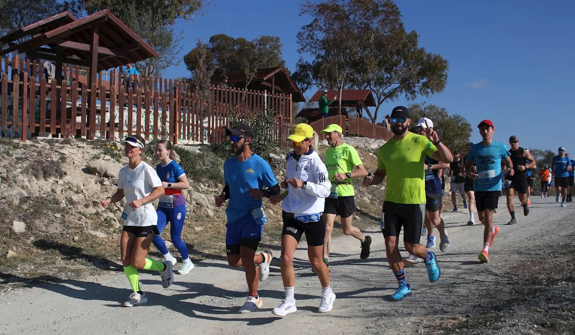 The image shows a group of people running outdoors, likely participating in a race or a fun run. They are wearing athletic clothing and running shoes, and the setting appears to be a rural or park area with trees and blue skies. Some participants are wearing caps and sunglasses, and there are wooden structures in the background.