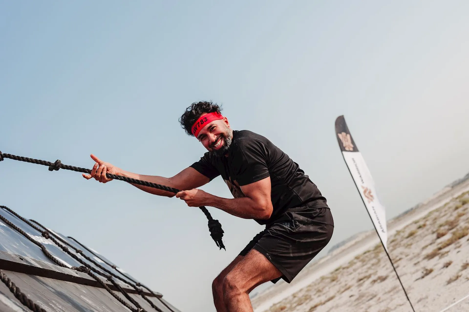 The image shows a person participating in some sort of outdoor obstacle course or physical challenge. The individual is wearing a red headband, a black t-shirt, and black shorts. He is smiling and appears to be pulling on a rope as part of the activity, with a focus on strength and endurance. In the background, there is a flat terrain that looks like it could be a beach or desert, and the sky is clear. Additionally, there is a banner or flag partially visible next to the person, which likely indicates a checkpoint or station within the event.