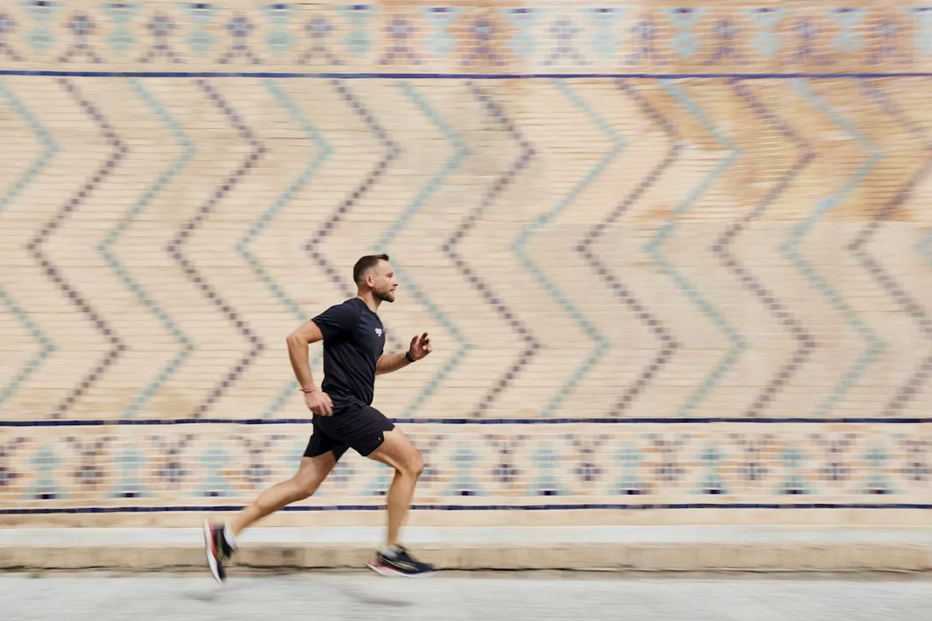The image shows a person running in front of a patterned wall. The wall has a zigzag design with various colors. The person is wearing athletic clothing, including a black T-shirt and shorts.