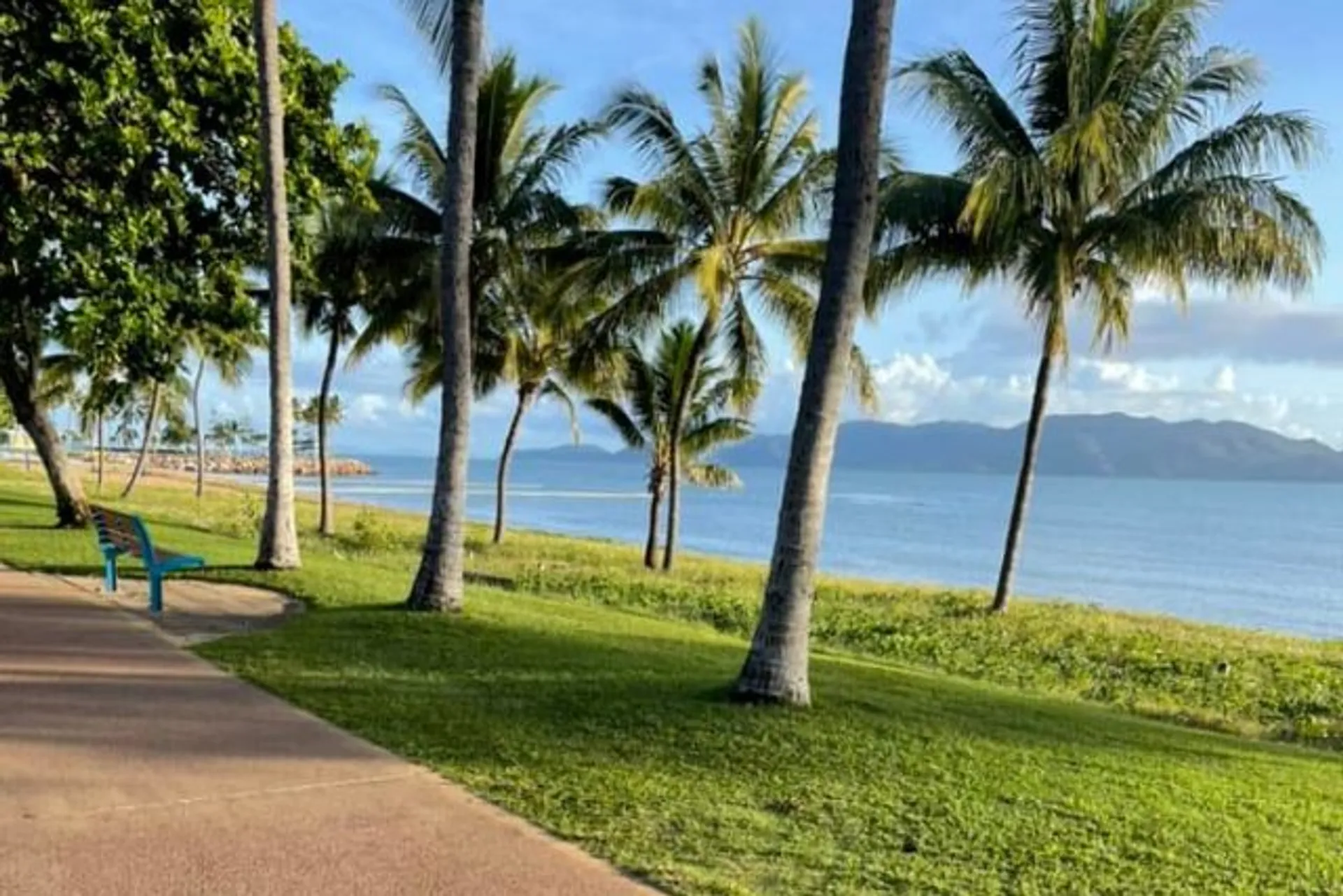 The image shows a scenic coastal landscape with tall palm trees lining a grassy area. A clear paved path or sidewalk meanders through the grass, inviting a leisurely stroll. To the right, there's a beautiful view of the sea with an island visible in the distance. The sky is partly cloudy, but plenty of blue sky is showing, suggesting a pleasant weather. A solitary blue bench faces the water, offering a place to sit and enjoy the serene environment. The overall scene is peaceful and picturesque, typical of a tropical or subtropical region.
