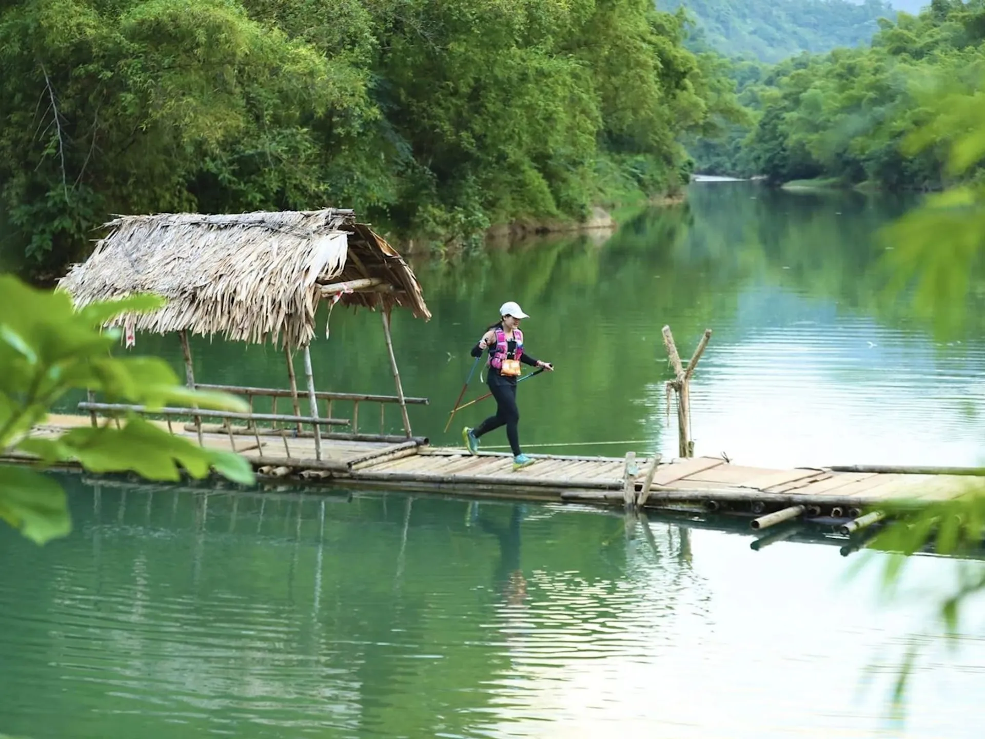The image shows a person running on a narrow bamboo bridge over a body of water. The bridge has a thatched roof shelter on one side. The setting is lush and green, with trees and vegetation surrounding the area, creating a serene and natural environment.
