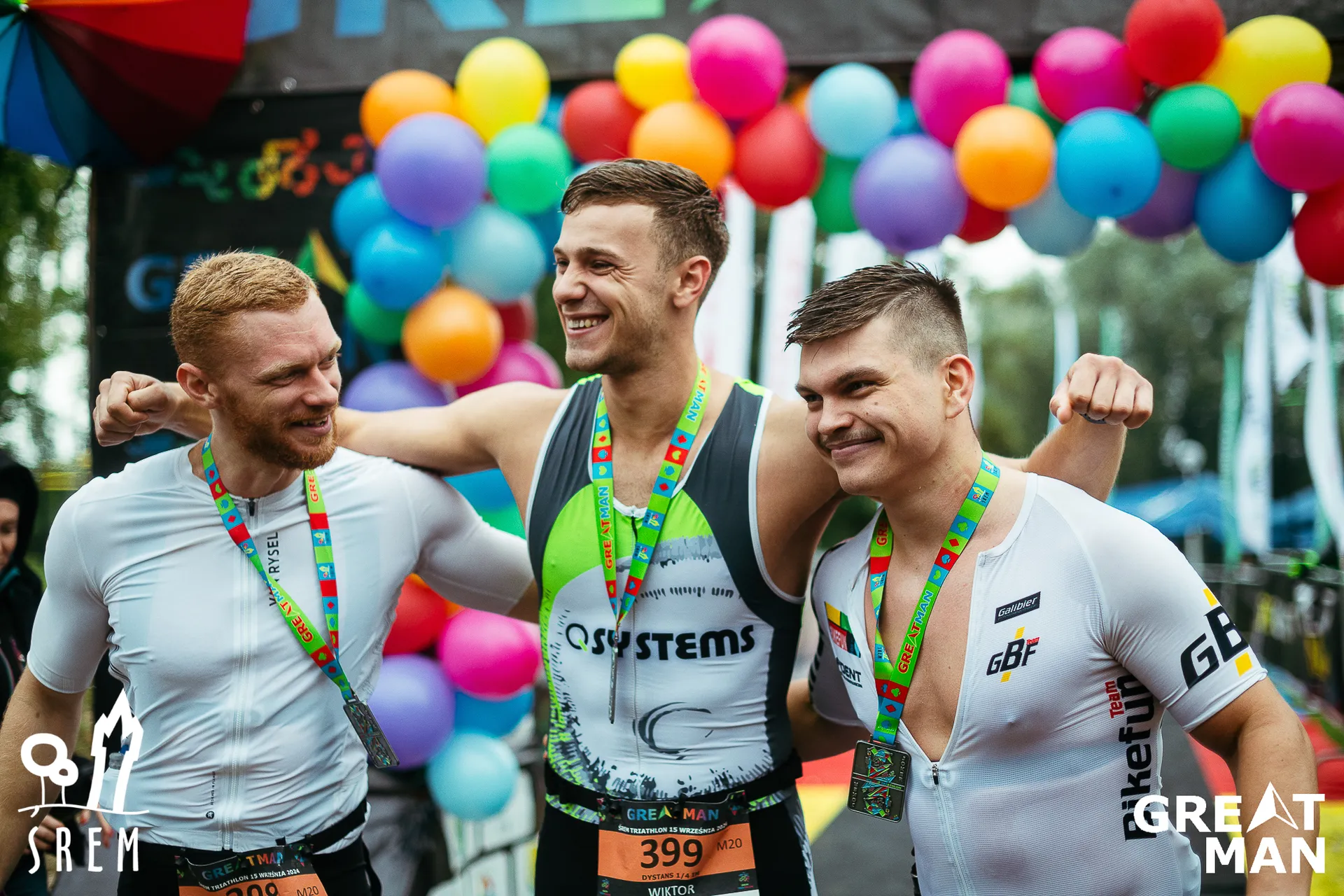 The image shows three athletes standing together, wearing race gear and finisher medals. They seem to be celebrating or posing for a photo after a sporting event, possibly a triathlon or a similar competition. There are colorful balloons in the background, and the athletes look happy. The event appears to have some branding with logos and text visible.
