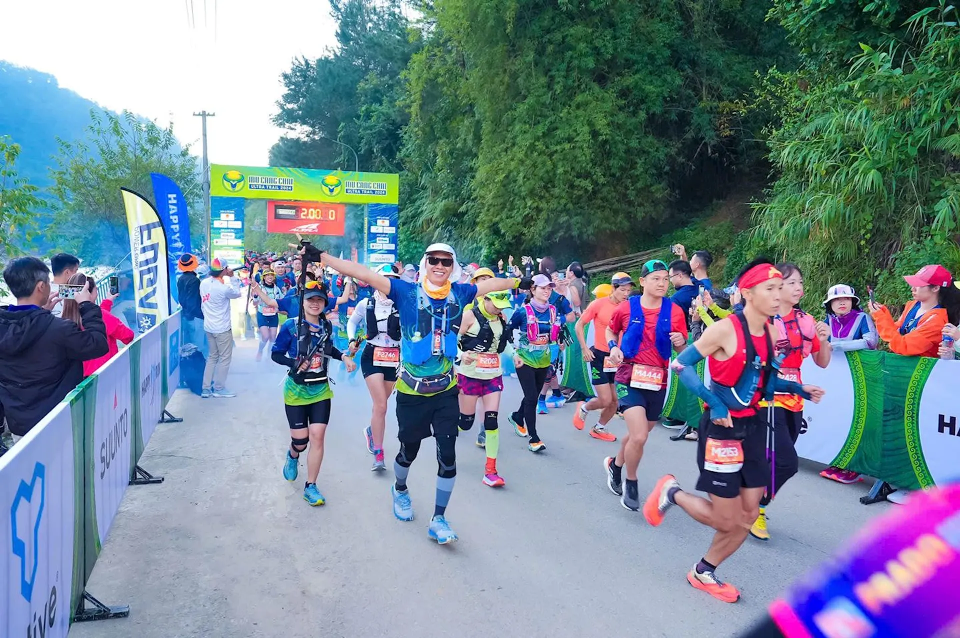 The image shows a group of runners participating in a marathon or running event. They are on a road lined with banners and spectators. The participants are wearing sports gear and race bibs. The background has trees, and there's a large event banner at the starting line.