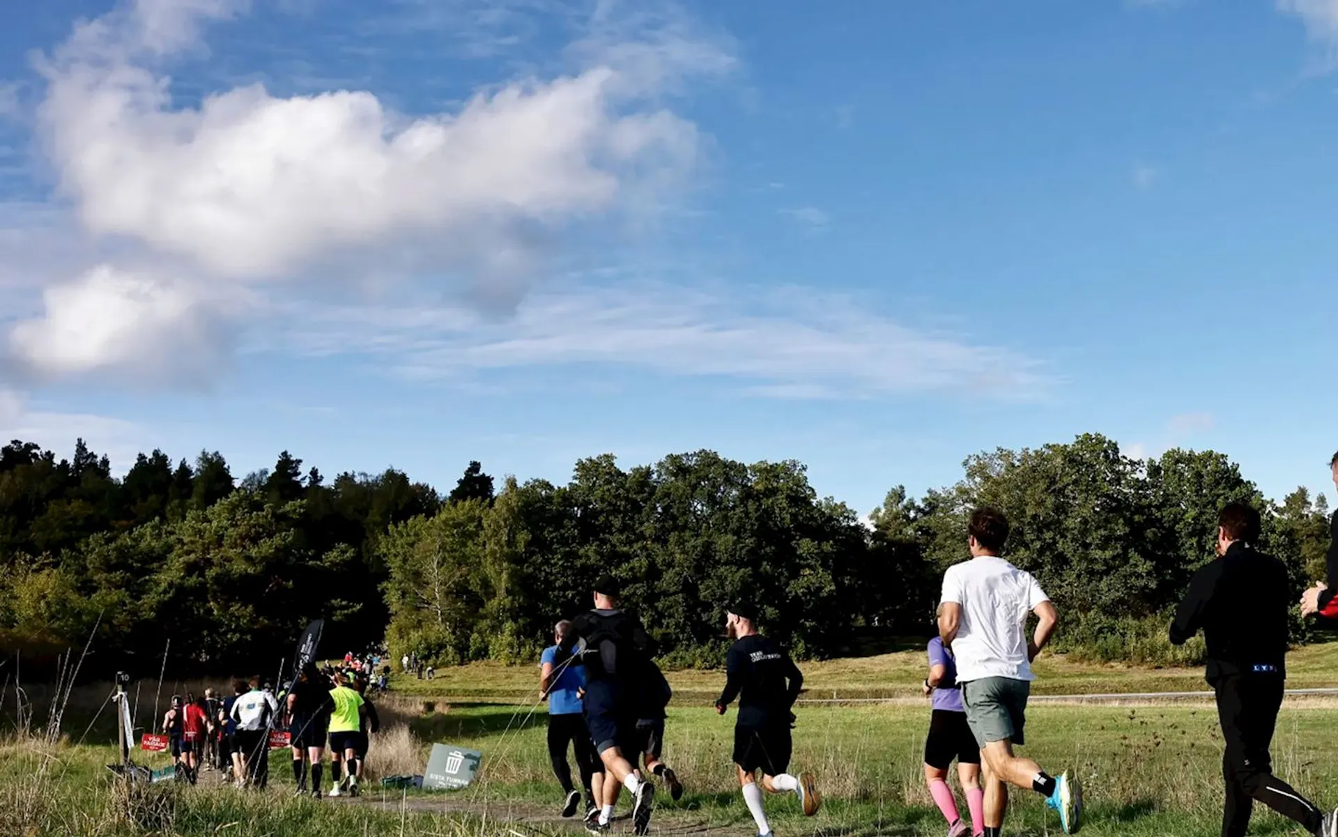 The image shows a group of people running outdoors on a grassy path or track. The scene is set in a natural environment with trees and a blue sky with scattered clouds. Some people are wearing athletic clothing and appear to be participating in a run or race.