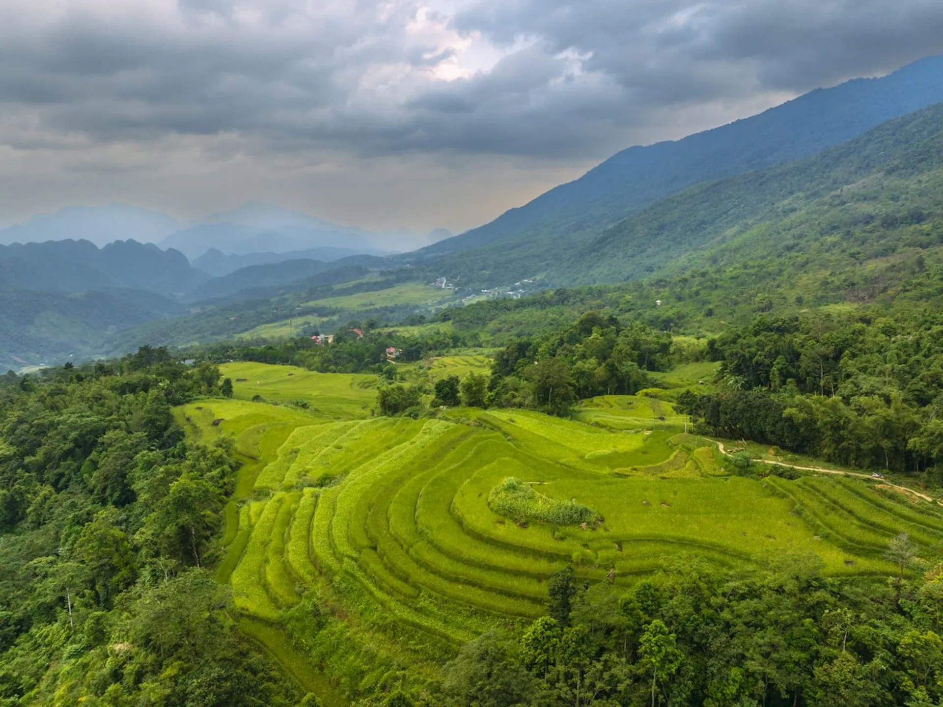 This image shows a scenic landscape with terraced rice fields on a hilly terrain. The fields are lush green, indicating they are likely in a growing season. In the background, there are mountains under a cloudy sky, creating a dramatic and picturesque view. The scene is likely rural and showcases natural beauty and agricultural practices.