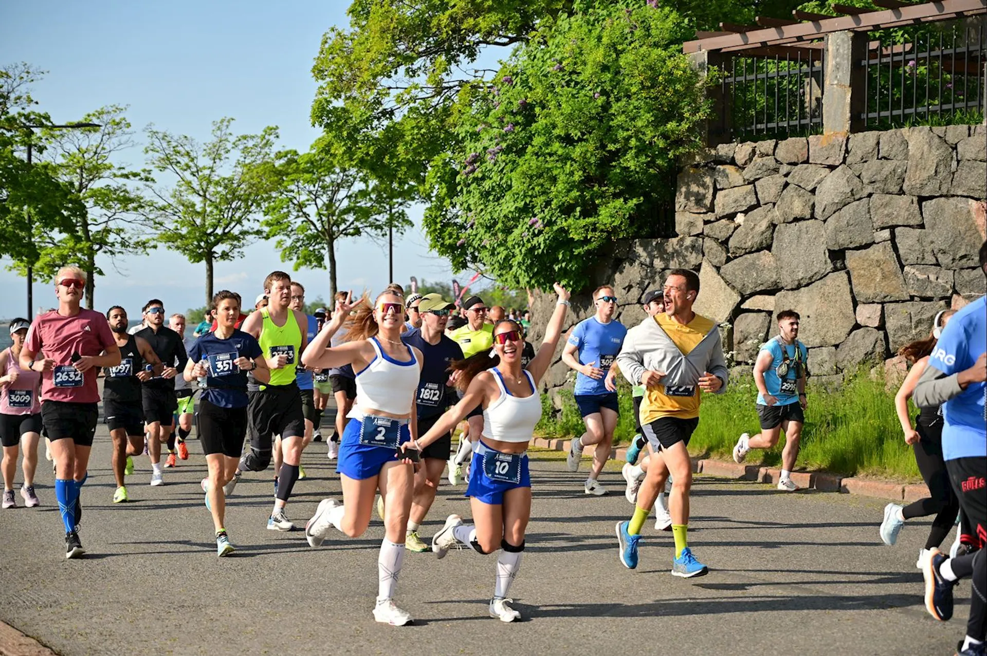 The image shows a group of people participating in a road race or marathon. They are running along a paved path lined with trees and a stone wall. The runners are wearing bib numbers and athletic clothing, and some are smiling and waving at the camera. The weather appears to be sunny and pleasant.