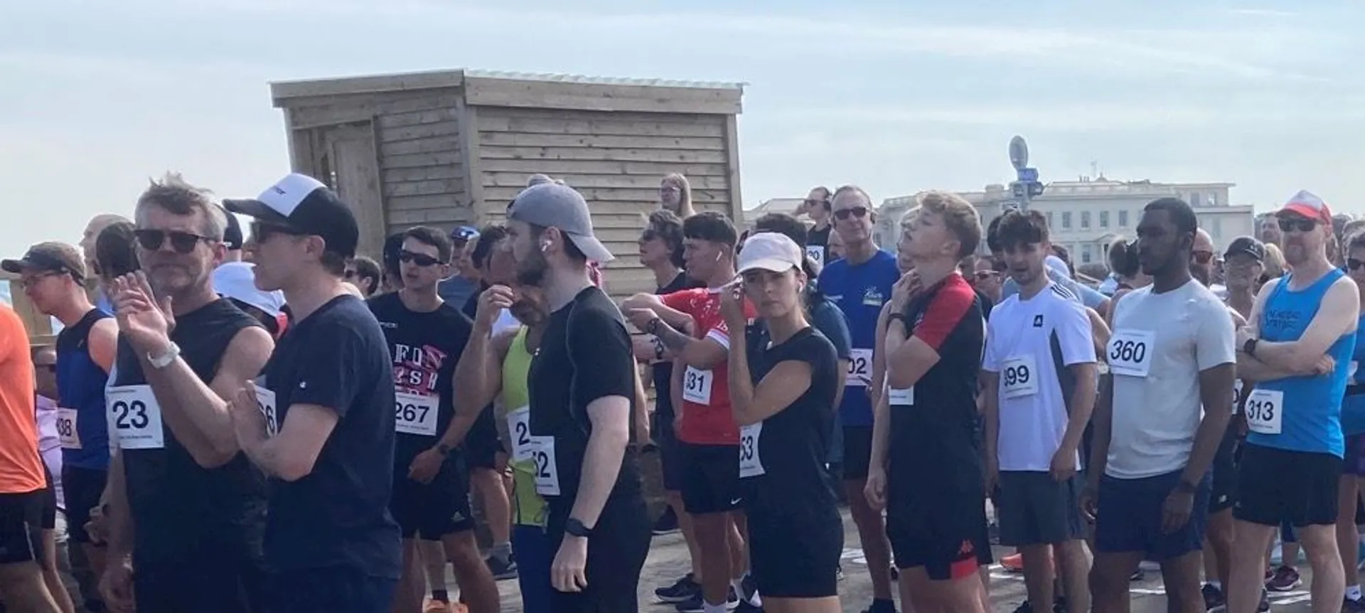 The image shows a group of people, many of whom are wearing athletic clothing, gathered together as if preparing for a race or a similar event. Some of the participants have numbered bibs attached to their shirts. The setting appears to be outdoors, possibly near a wooden structure and some buildings in the background.