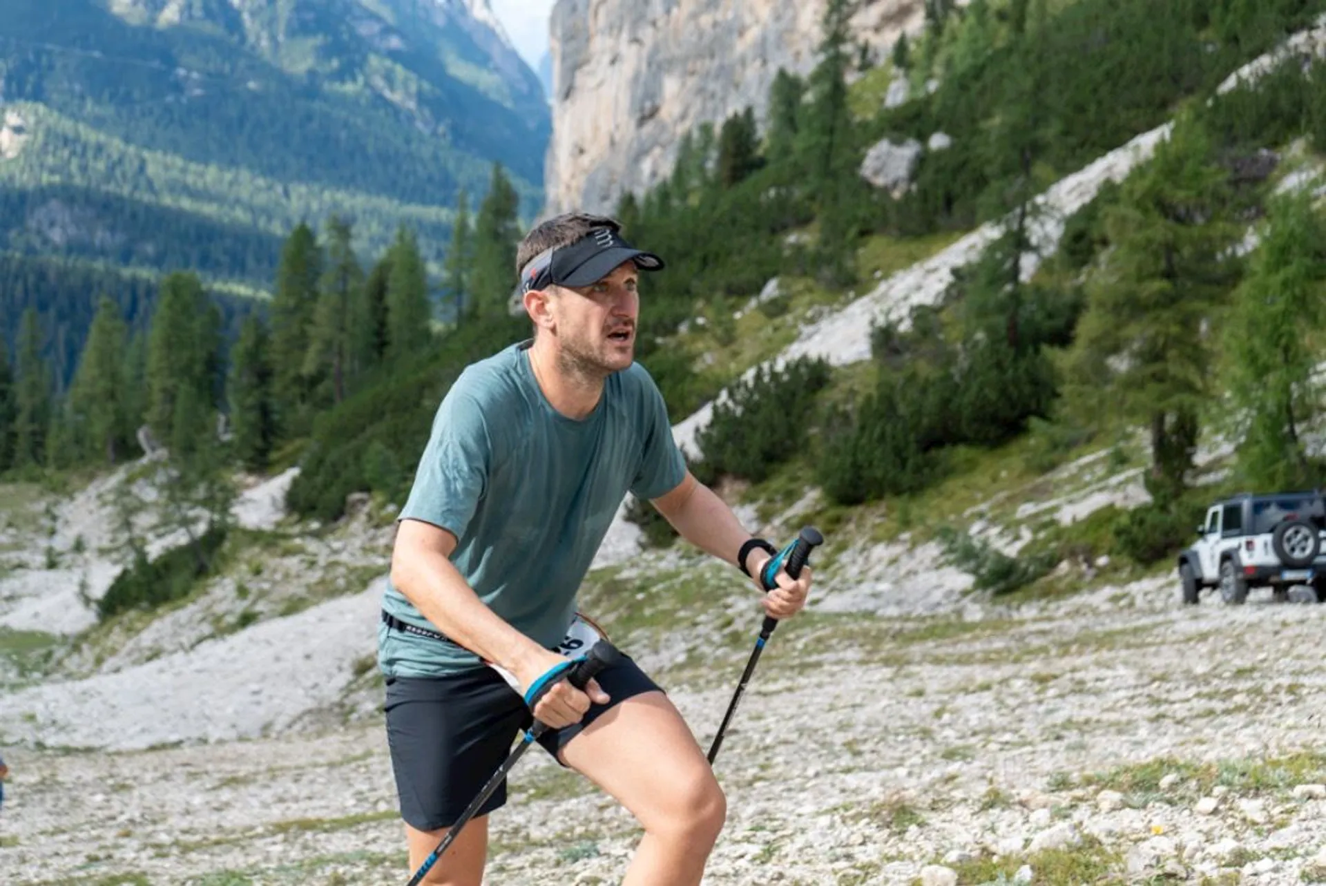 The image shows a man engaged in an outdoor activity, possibly hiking or trail running, in a mountainous area. He's equipped with trekking poles, suggesting that he might be on a challenging terrain. He's wearing a t-shirt, shorts, a cap, and a watch — typical gear for an outdoor sports enthusiast. Behind him, there's a rugged landscape with trees and steep mountain slopes, indicative of an alpine region. Additionally, there is a vehicle, which looks like an off-road SUV, parked on a gravel area in the background, which could suggest the proximity of a road or a starting point for a trail. The man appears to be in motion, focused on his activity, and possibly navigating across rocky ground.