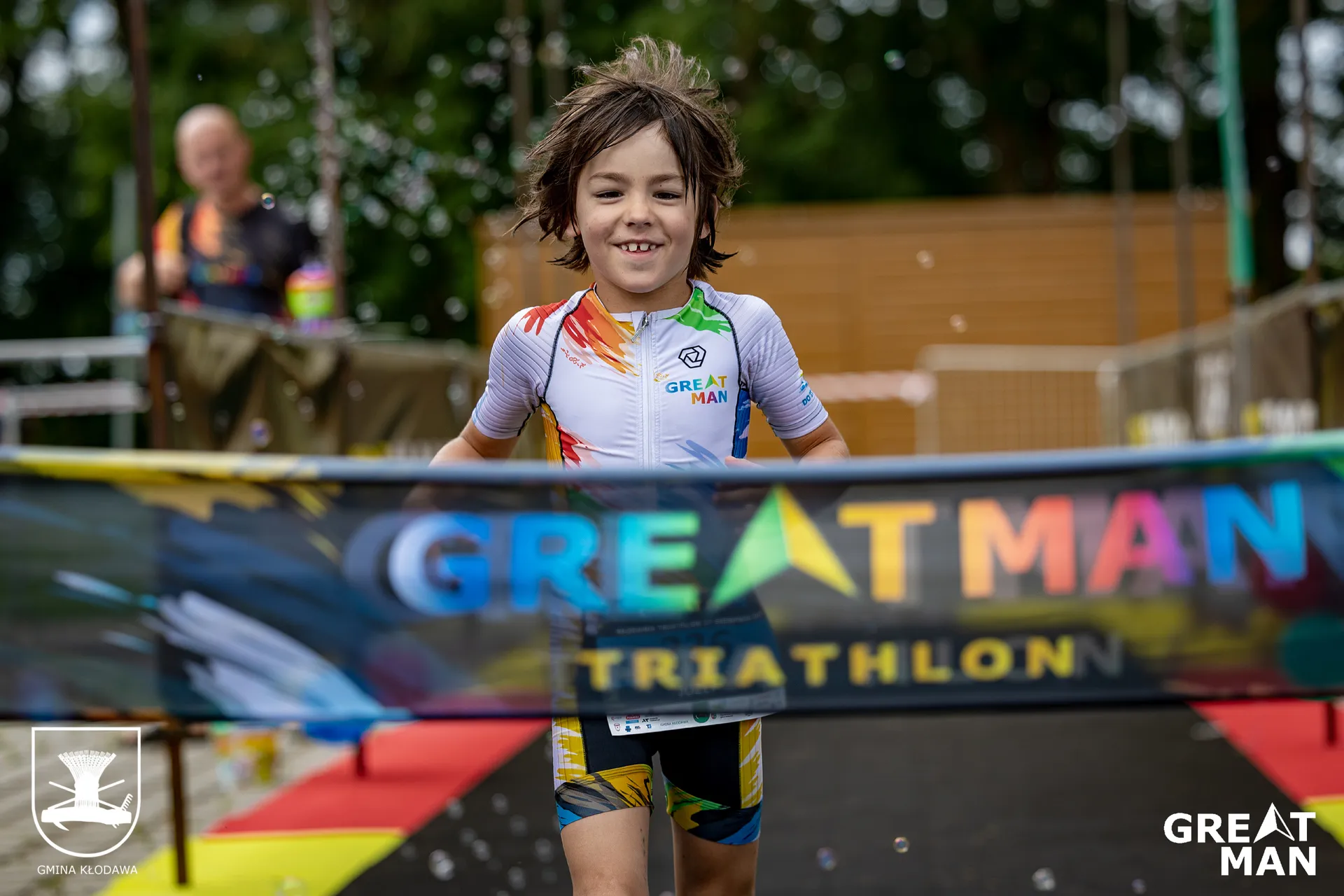 The image shows a young participant crossing the finish line at the "Great Man Triathlon." The child is wearing a colorful triathlon suit with event branding. There are visible details such as bubbles and the finish line ribbon.