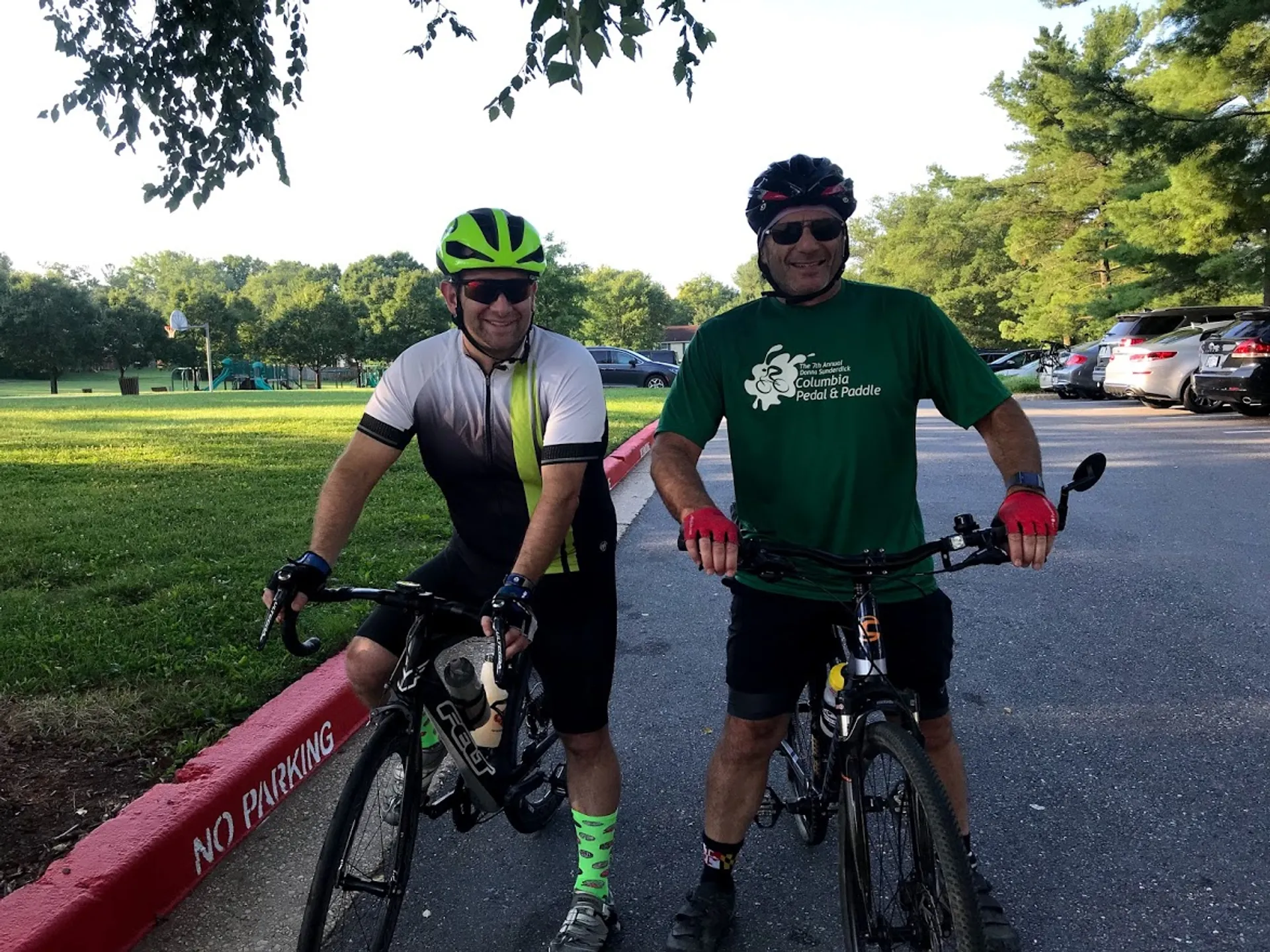 The image shows two individuals standing outdoors beside bicycles. They both appear to be dressed in cycling attire, with one wearing a bright green helmet and the other a black one. They are smiling and seem to be enjoying themselves. The surrounding environment suggests that they might be in a park or a recreational area, as there is green grass in the background and a "NO PARKING" sign is visible painted on the curb. The time of day appears to be either morning or evening based on the lighting and shadows.