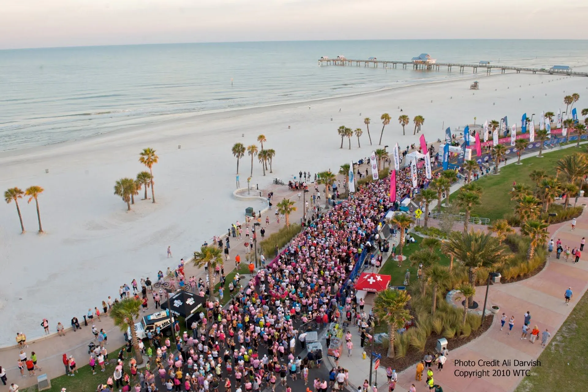 The image shows a large crowd of people gathered for an event by a beachfront