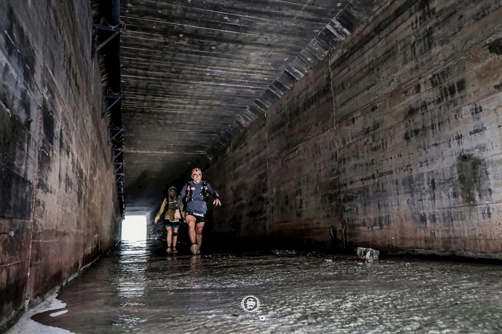This image shows two people walking through a large drainage tunnel or culvert. The tunnel has concrete walls and a shallow stream of water flowing along its base. The people appear to be geared in outdoor or hiking attire, likely exploring or traversing through the tunnel. The scene is lit by natural light coming from the end of the tunnel.