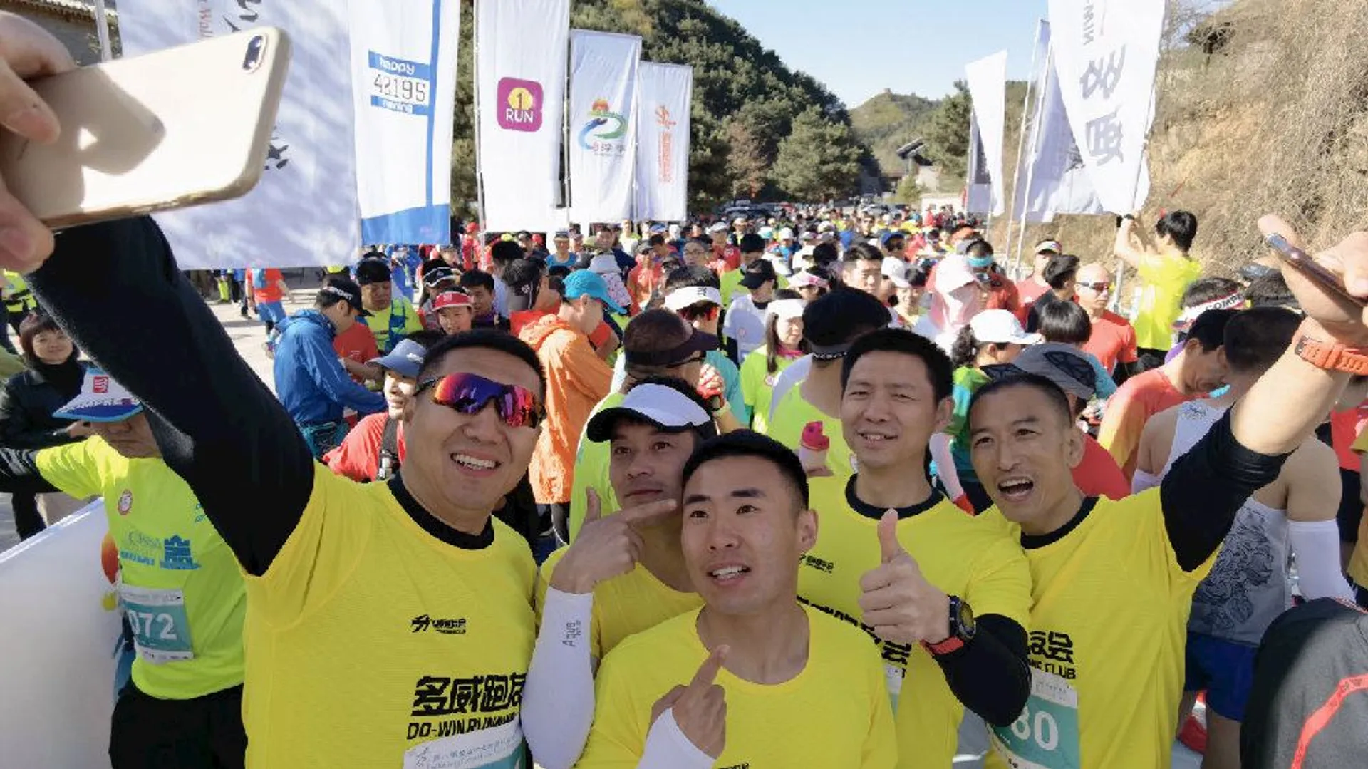 The image shows a group of people participating in a running event, most likely a marathon or race based on the racing bibs visible on their shirts. A few individuals in the foreground are posing for a selfie, with one person holding a smartphone camera to capture the moment. They are all wearing matching yellow shirts, which could suggest they are part of a team or club. The runners are wearing various athletic gear suitable for a race. In the background, numerous other participants and event banners are visible, contributing to the lively atmosphere of the race. Everyone seems to be in high spirits, as indicated by the smiles and thumbs-up gesture from one of the runners.