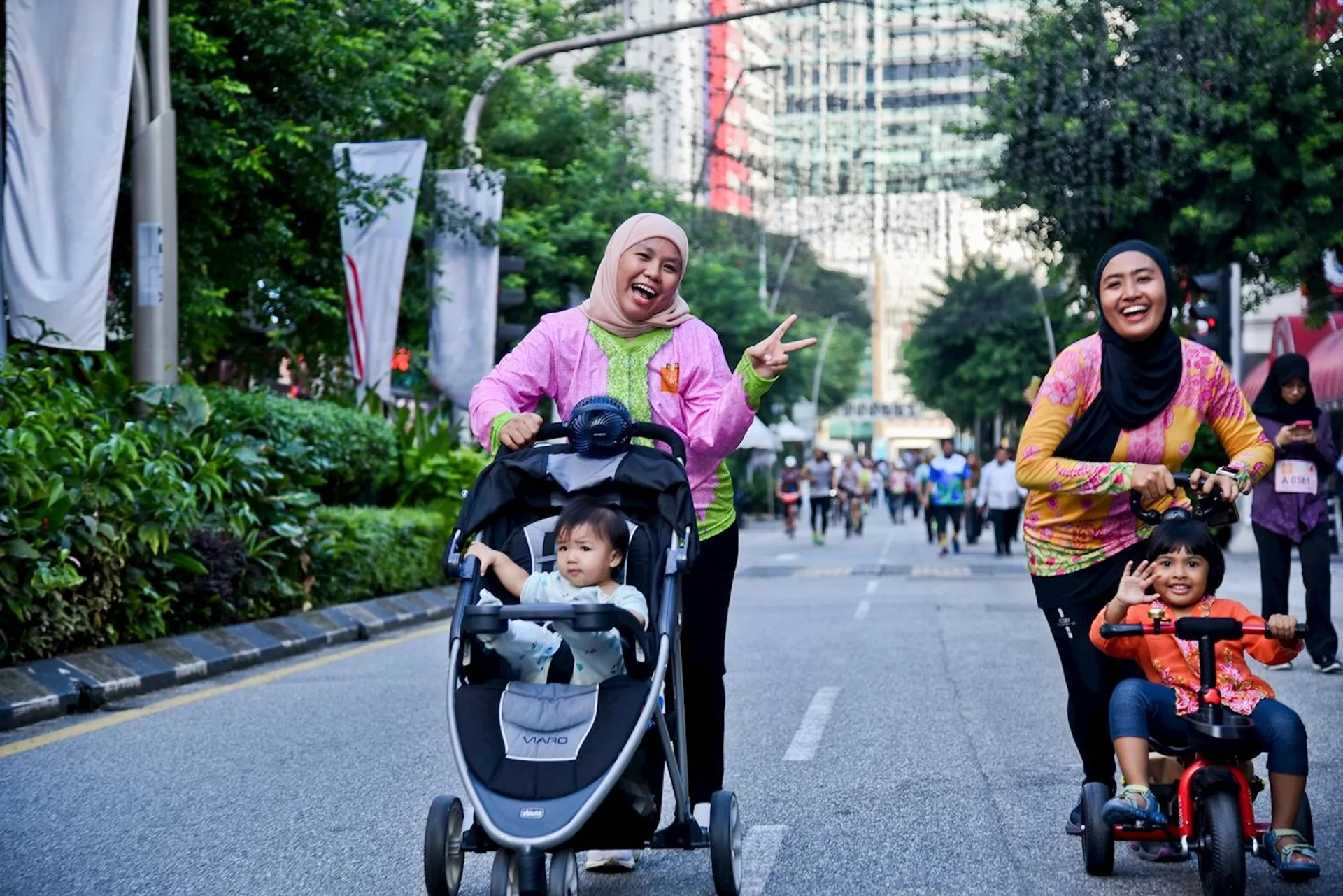 The image shows two women and two young children on a street. One woman is pushing a child in a stroller and giving a peace sign, while the other woman is smiling with a child on a small bicycle. There are more people walking in the background. The scene is outdoors, possibly during a public event or walk.
