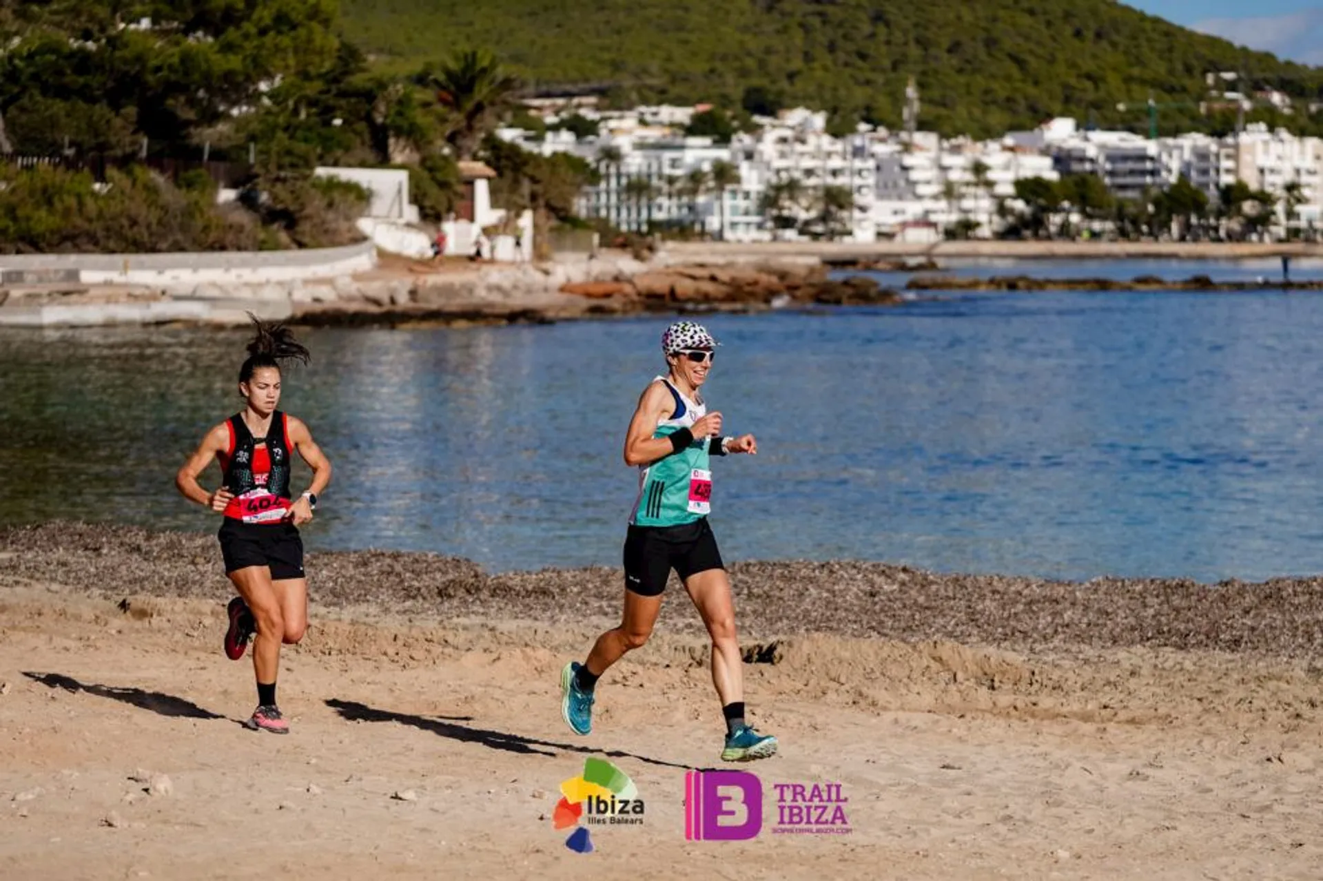 This image shows two people running along a beach. They're wearing athletic gear, including race bibs. The background features water, trees, and a distant shoreline with buildings. There are logos for "Ibiza Marathon" and "Trail Ibiza" in the foreground.