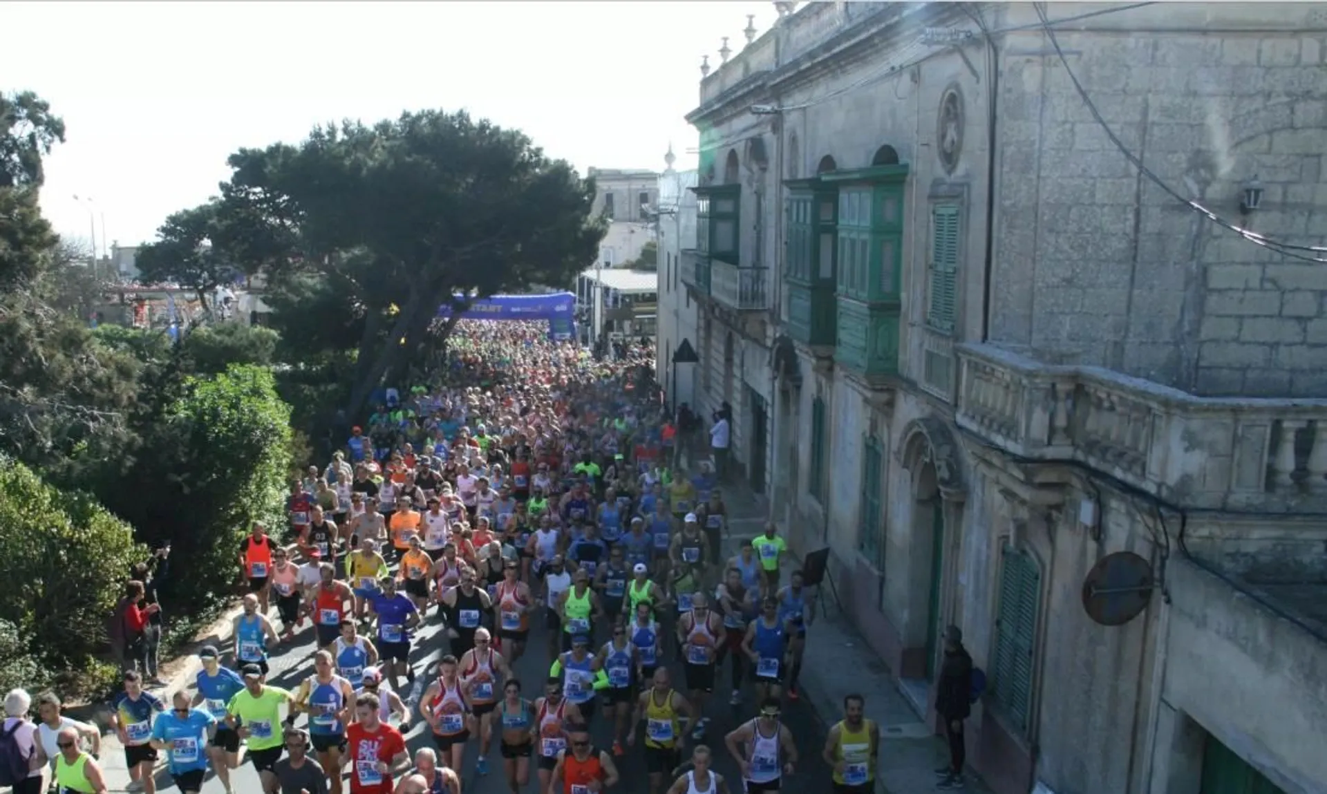 The image shows a large group of runners participating in a road race. They are running along a street lined with buildings and greenery. There's a banner in the background, potentially indicating the start or finish line of the race. The participants are wearing athletic clothing and number bibs.