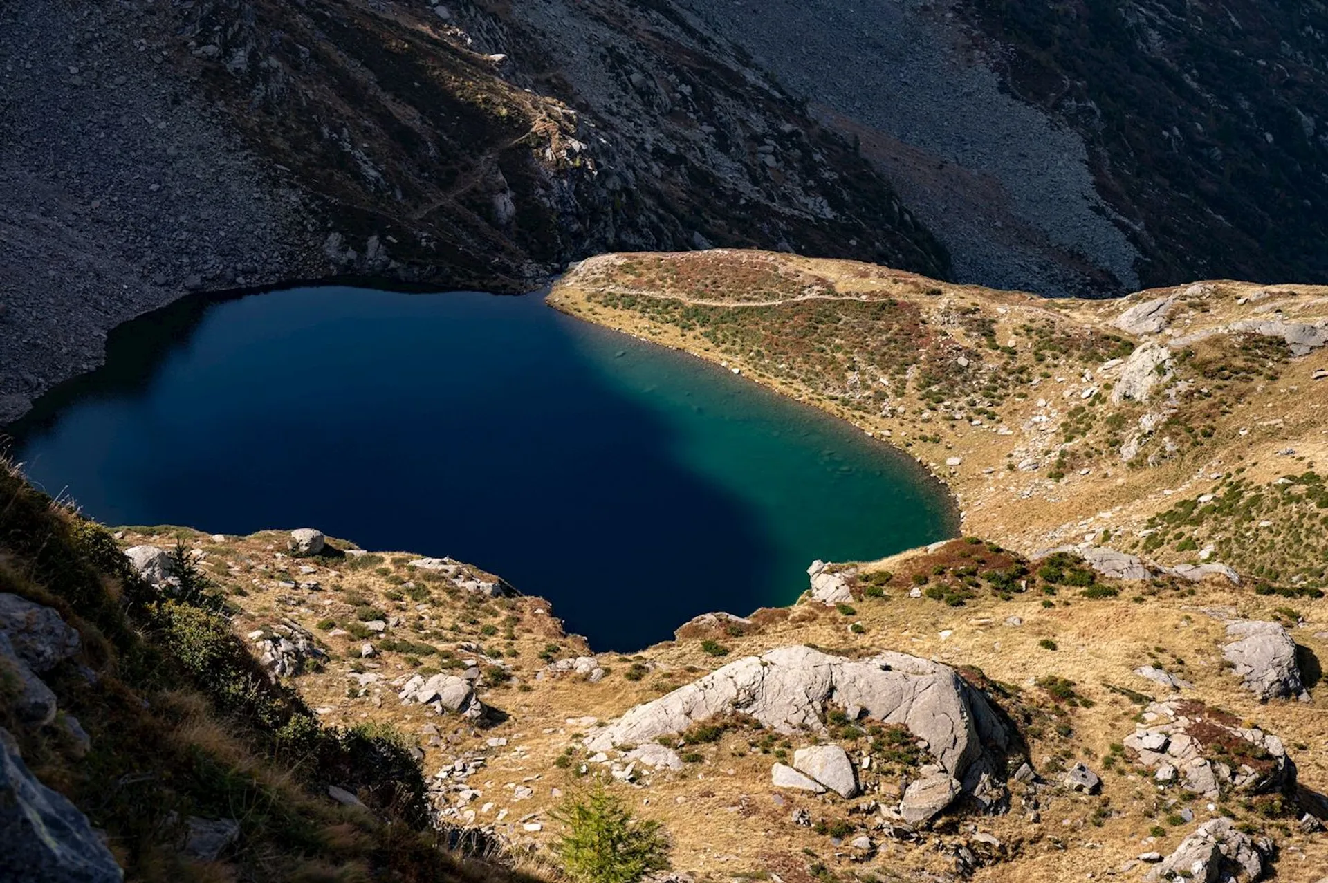 This image shows a lake surrounded by rocky and grassy terrain. The water appears dark blue, and there are slopes with scattered vegetation leading down to the lake. The landscape suggests a mountainous or high-altitude region.