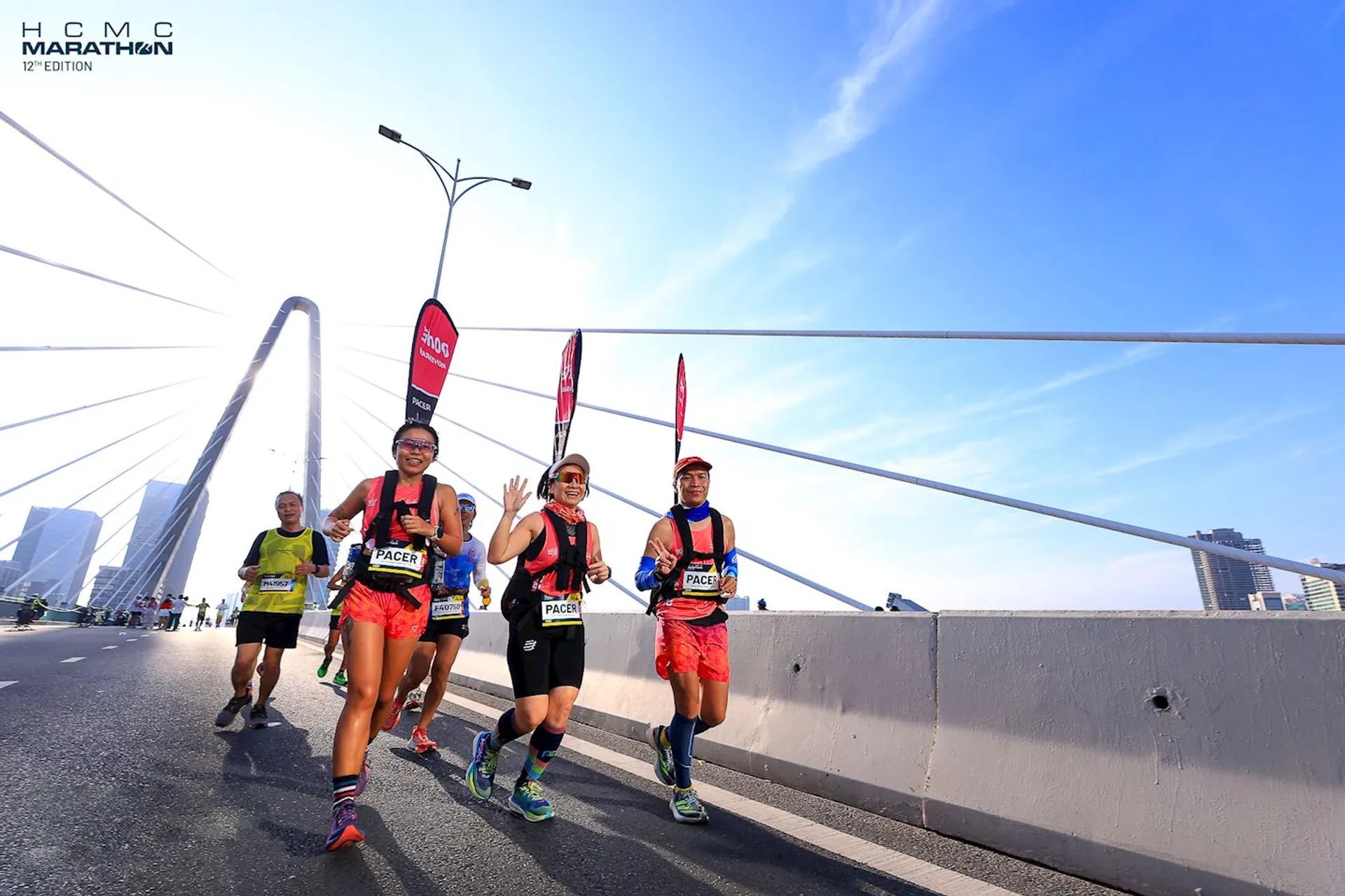 The image shows a group of marathon runners on a bridge during a race. They are wearing athletic gear, and some have "PACE" written on their outfits, indicating they are likely pacers for the event. There are marathon banners and signs visible, and the bridge's architecture is prominent in the background. The setting appears to be during the day under clear skies.