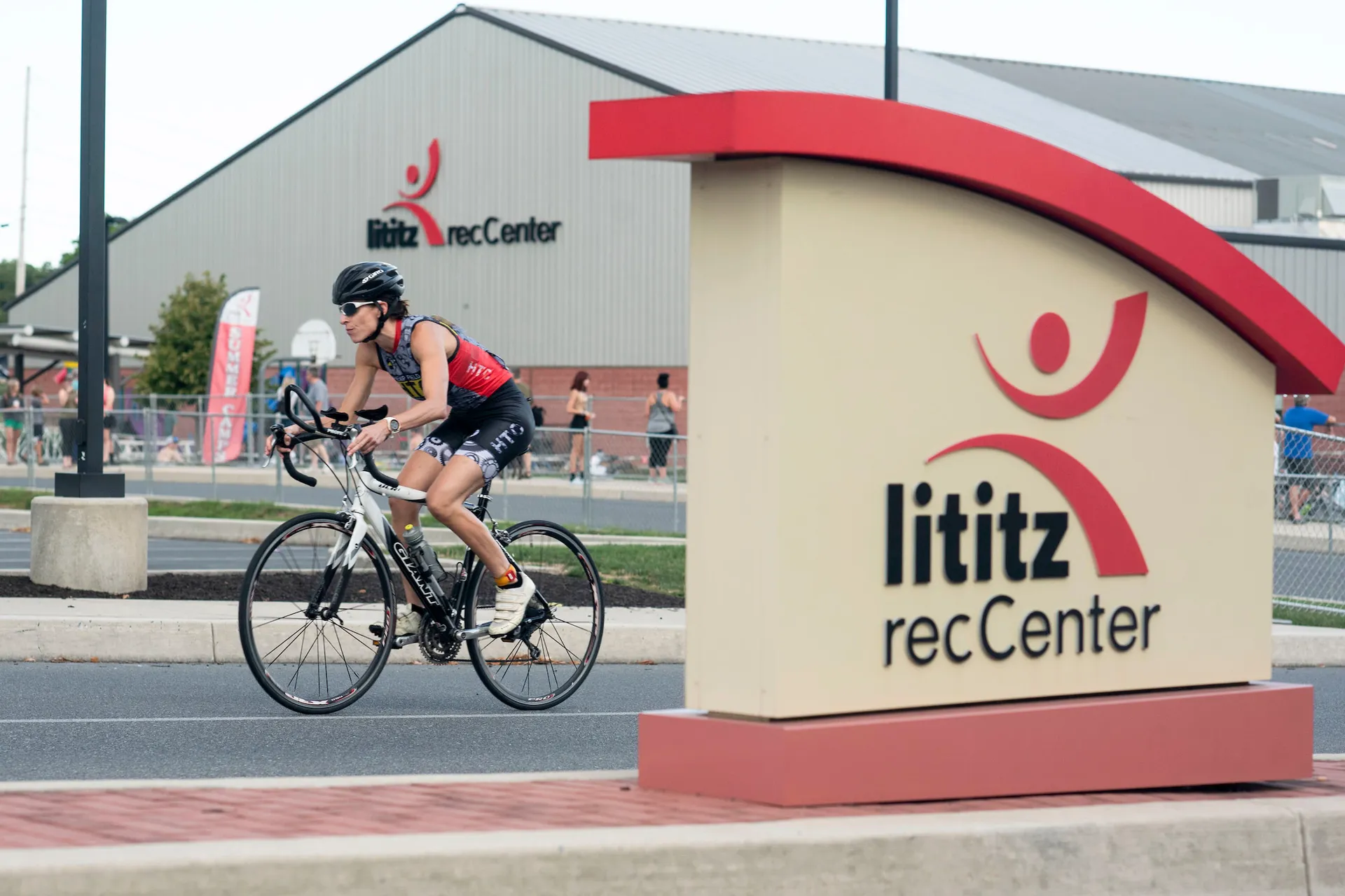 In the image, there's a cyclist wearing a helmet and sports attire—red and black—riding a road bike on a paved street or pathway. Behind the cyclist, there's a building with a sign that reads "Lititz recCenter," indicating that the scene likely takes place near a recreational facility in the town of Lititz. The center's logo, which features an abstract figure with arms raised, is also visible on the sign in the foreground. The setting appears to be in a developed area with a focus on community and outdoor activities.