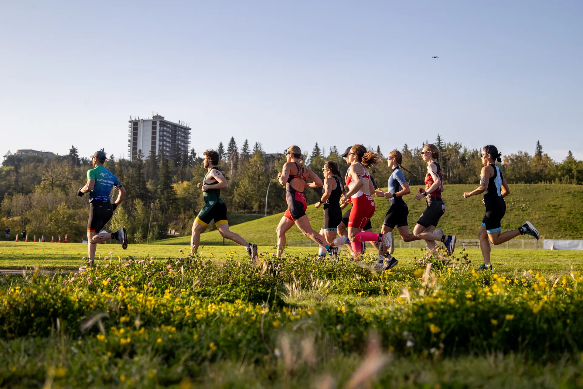 In this image, there is a group of people engaged in a running activity, likely a race or a group run. The setting appears to be a grassy area with wildflowers and trees, suggesting it might be a park or a similar outdoor space. There's a clear sky, and the sunlight is casting shadows, indicating that the photo was taken on a sunny day. In the background, there are buildings that suggest the location is near an urban area. Additionally, there is a flying object in the sky, which could possibly be a bird or a small drone, but it's too distant to be identified clearly. Everyone is wearing athletic gear, emphasizing the physical activity context of the scene.