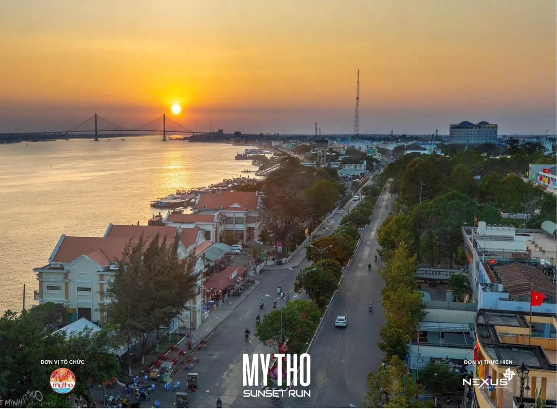 The image depicts a scenic view of a city at sunset. In the foreground, we see a tree-lined street with buildings that have a mix of traditional and modern architecture. On the left, there is a wide river with a prominent bridge spanning across it, with the setting sun creating a golden hue on the horizon. The sky is painted with warm tones of orange and yellow, blending into a light blue.

The city appears to be lively and bustling, with vehicles on the roads and a few visible pedestrians. Along the river, there are boats which might be used for transportation or fishing. There's a large antenna tower visible in the background.

In the top right corner of the photo, there is text "MYTHO SUNSET RUN" along with