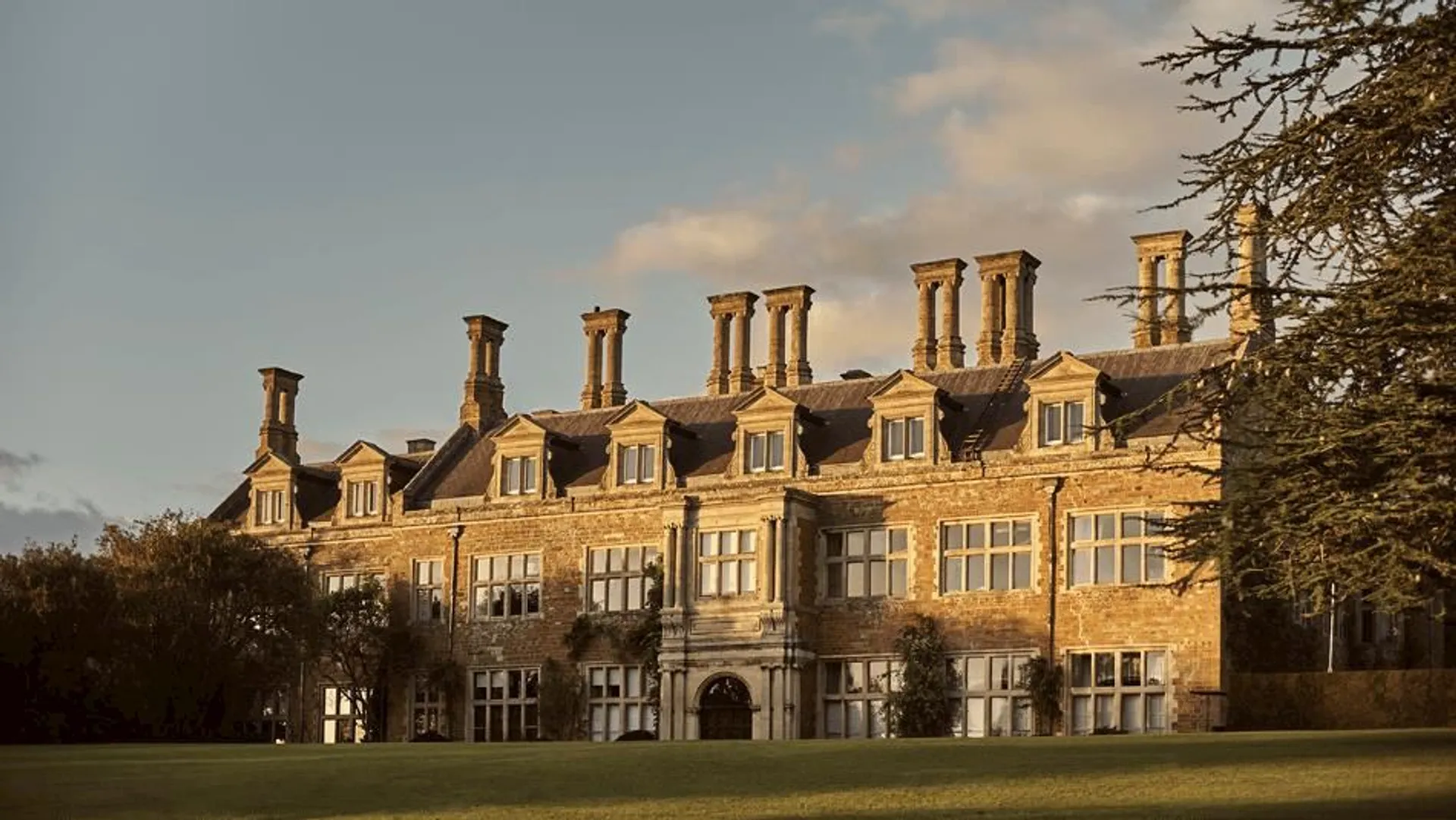 The image shows an old, large building, likely a historical mansion or estate. It has multiple chimneys, a stone exterior, and several windows. The architecture suggests it might be from a past era, with a classic and grand appearance. In the foreground, there is well-maintained grass, and there are trees around the building. The lighting suggests it might be late afternoon or early evening.