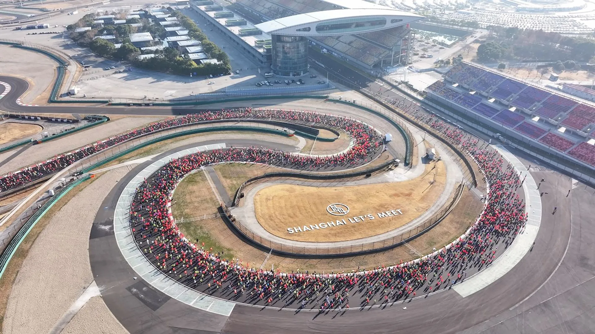 The image shows an aerial view of a race track, specifically Turn 1 at the Shanghai International Circuit. The track has a unique, spiral shape, and there are a large number of people walking or participating in an event on it. The grandstand and surrounding facilities are visible, along with some vehicles. The ground has text that says "SHANGHAI LET’S MEET.”