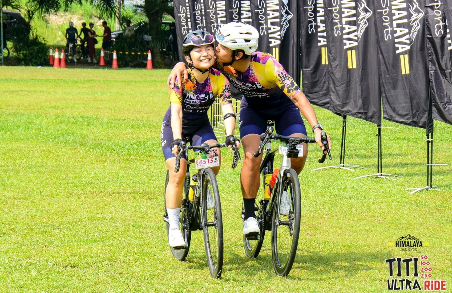 The image shows two cyclists on a grassy field during a cycling event. They are both smiling and one is giving the other a kiss on the cheek while riding. Both wear helmets and matching cycling outfits. There are banners in the background that say "Himalaya Sports" and the event name "Titi Ultra Ride" is visible in the bottom right corner.