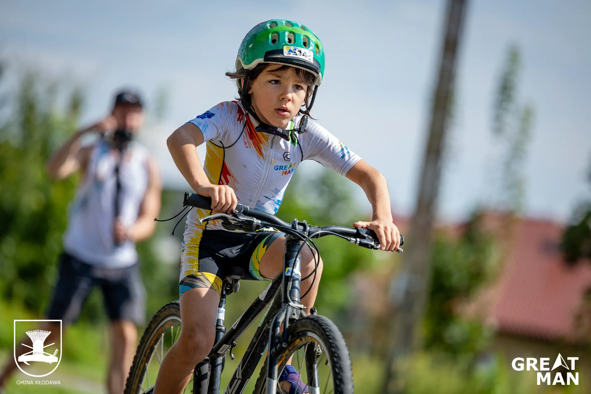 The image shows a child wearing a helmet cycling outdoors. The child is dressed in cycling gear and appears to be focused on riding. There's a blurry figure with a camera in the background, suggesting the setting might be part of an event or race. The logos in the corners seem to be from an organization or event called "Great Man."