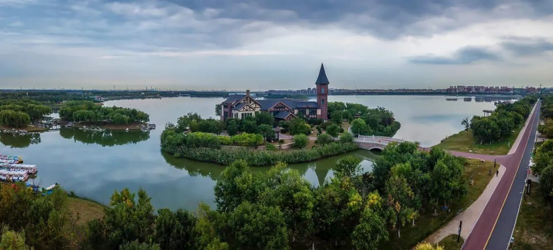 The image shows a scenic view of a small island with a building featuring a tower, surrounded by a body of water. The area is lush with trees and greenery. There's a bridge connecting the island to the mainland. In the background, a road and more trees are visible, and the sky is partly cloudy.